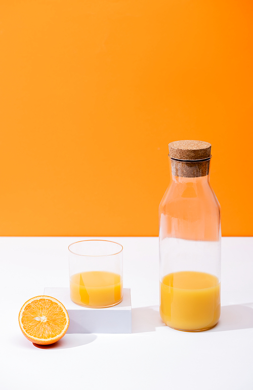 fresh orange juice in glass and bottle with cork near cut fruit on white surface isolated on orange