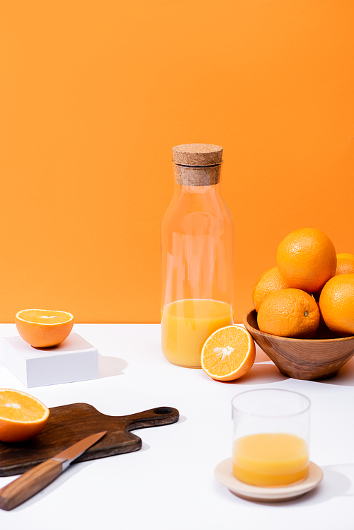 fresh orange juice in glass and bottle near oranges in bowl, wooden cutting board with knife on white surface isolated on orange