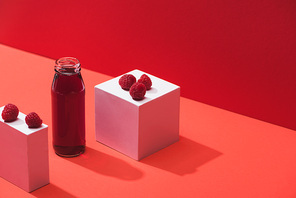 fresh berry juice in glass bottle near ripe raspberries on cubes on red background