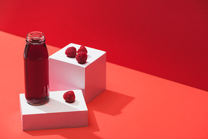 fresh berry juice in glass bottle near ripe raspberries on cubes on red background