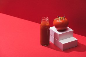 fresh vegetable juice in glass bottle near ripe tomato on stand on red background