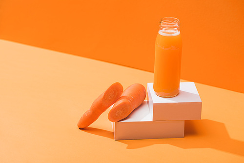 fresh juice in glass bottle near ripe carrots on cubes on orange background