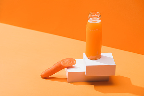 fresh juice in glass bottle near ripe carrot on cubes on orange background