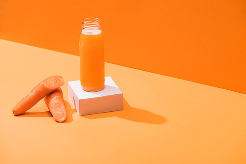 fresh juice in glass bottle on cube near ripe carrots on orange background