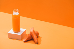 fresh juice in glass bottle on cube near ripe carrots on orange background