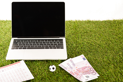 toy soccer ball, euro banknotes, betting lists near laptop with blank screen on green grass isolated on white, sports betting concept