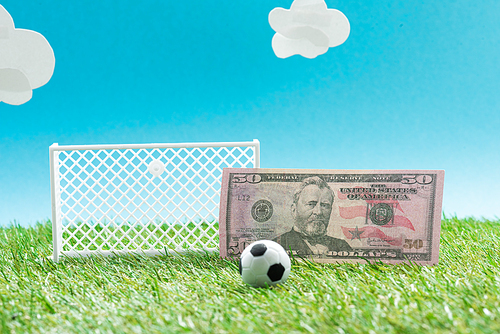 toy soccer ball and gates near dollar banknote on blue background with clouds, sports betting concept