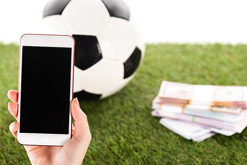 partial view of female hand with smartphone near packs of money and soccer ball on green grass isolated on white, sports betting concept