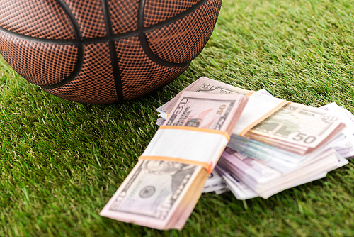 close up view of basketball ball near dollar and euro banknotes on green grass, sports betting concept