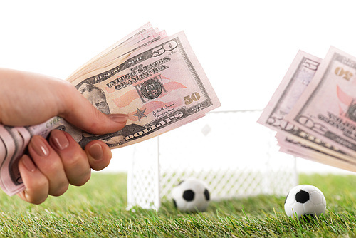 cropped view of woman holding dollar banknotes near toy football gates and balls isolated on white, sports betting concept