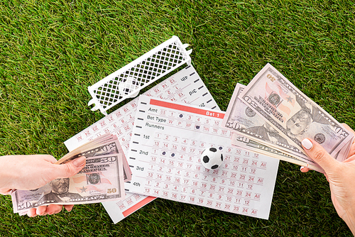 partial view of female hands with dollar banknotes near toy soccer balls, gates and betting lists on green grass, sports betting concept