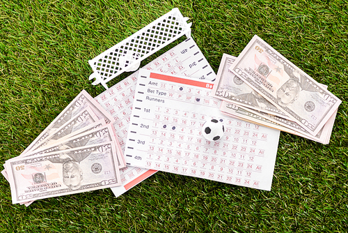 top view of toy football gates and ball near dollar banknotes and betting lists on green grass, sports betting concept