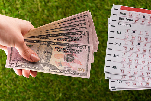 cropped view of woman holding dollar banknotes near betting lists on green grass, sports betting concept
