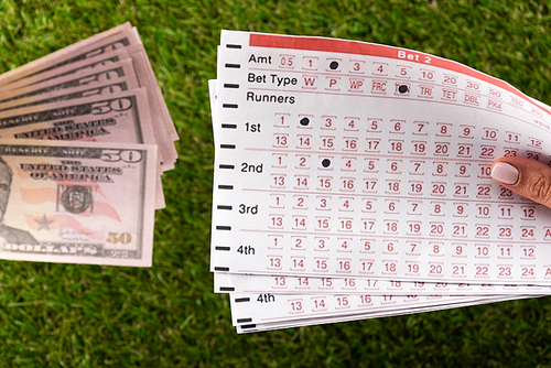cropped view of female hand with betting lists near dollar banknotes on green grass, sports betting concept