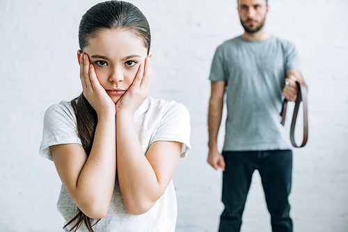 cropped view of father holding belt and upset daughter at home