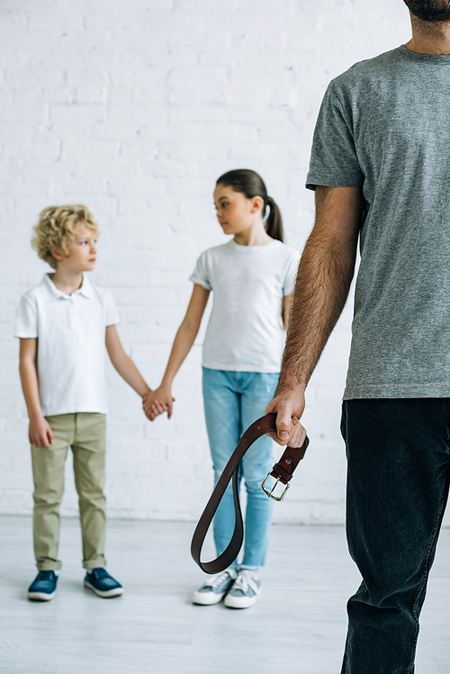 partial view of abusive father with belt and kids holding hands