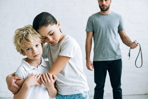 partial view of abusive father with belt and embracing kids at home