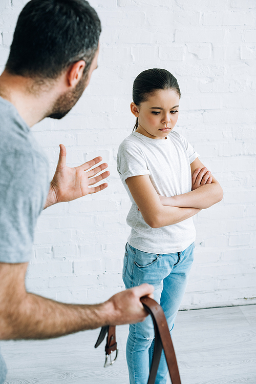 partial view of father holding belt and scolding upset daughter at home