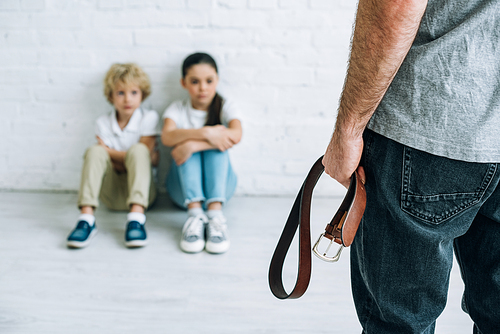 cropped view of abusive father holding belt and sad kids sitting on floor