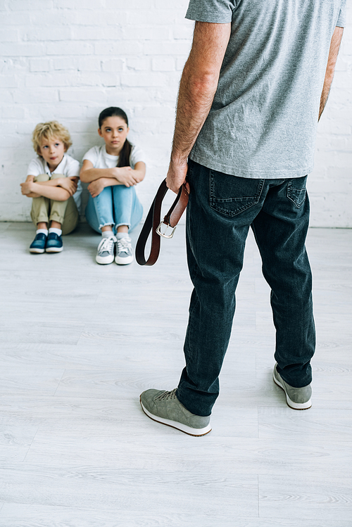 cropped view of abusive father holding belt and sad kids sitting on floor