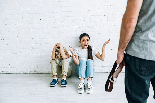 cropped view of abusive father holding belt and scared kids sitting on floor