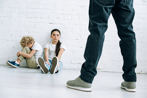cropped view of abusive father holding belt and sad kids sitting on floor