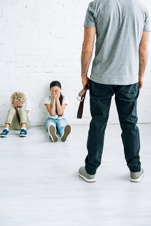 cropped view of abusive father holding belt and scared kids sitting on floor