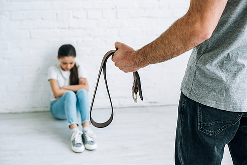 cropped view of father holding belt and sad daughter sitting on floor