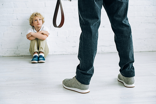 partial view of abusive father holding belt and son sitting on floor