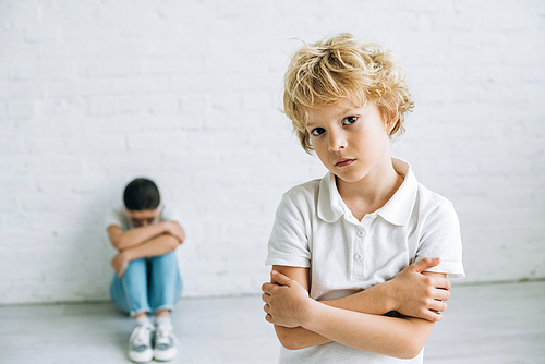 sad boy standing with crossed arms while sister sitting on floor and crying at home