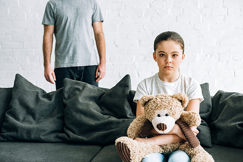 partial view of father and sad daughter with teddy bear sitting on sofa
