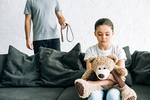 partial view of abusive father with belt and sad daughter with teddy bear sitting on sofa