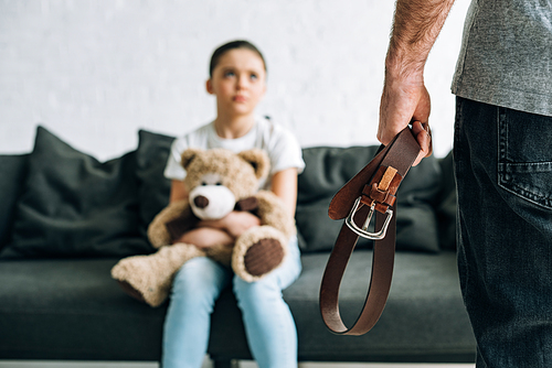 partial view of abusive father holding belt and scared daughter with teddy bear sitting on sofa