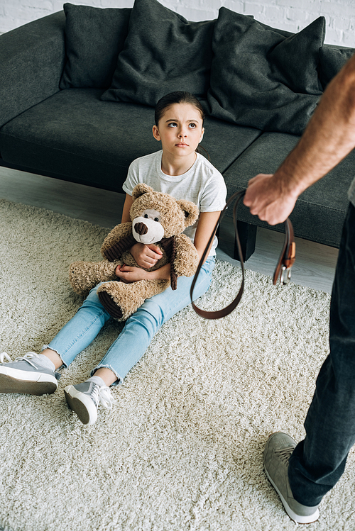 overhead view of abusive father holding belt and scared daughter with teddy bear sitting on carpet