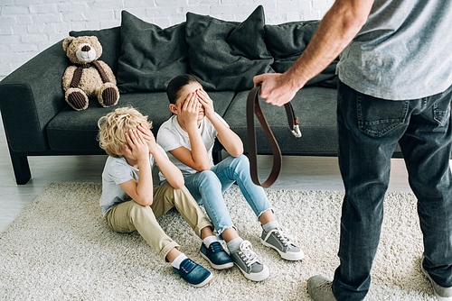 partial view of father with belt and upset kids sitting on floor