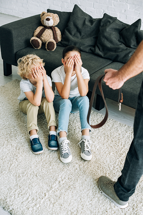 partial view of father with belt and upset kids sitting on floor