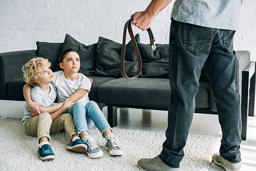 partial view of father with belt and upset kids sitting on floor