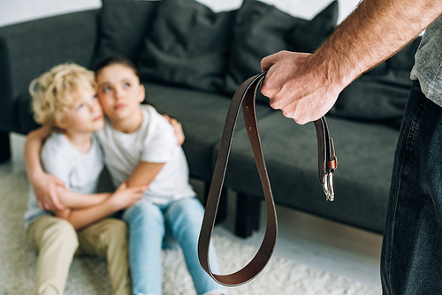 partial view of father with belt and upset kids sitting on floor