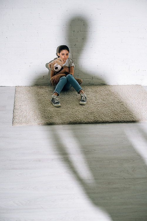 scared child with teddy bear sitting on carpet and big silhouette