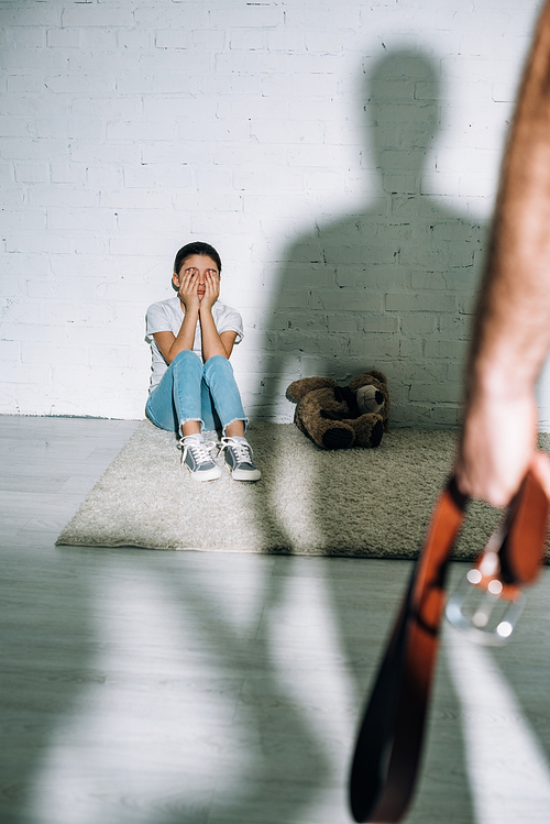 cropped view of father holding belt and scared daughter sitting on carpet near his silhouette