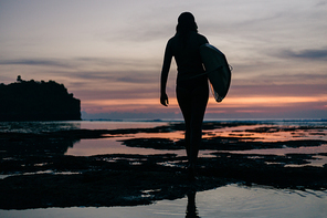 rear view of silhouette of woman with surfboard walking by seashore in evening