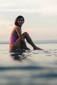 young girl sitting on surfboard in water in ocean at sunset