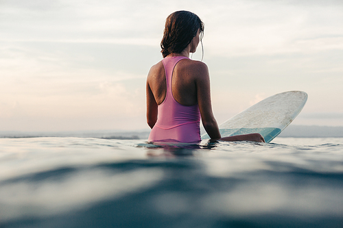 rear view of sportswoman sitting on surfboard in water in ocean at sunset