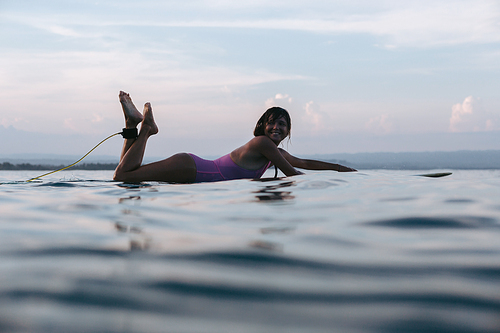 smiling woman lying on surfboard in water in ocean at sunset
