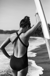 back view of young girl in swimsuit posing with surfboard near ocean, black and white