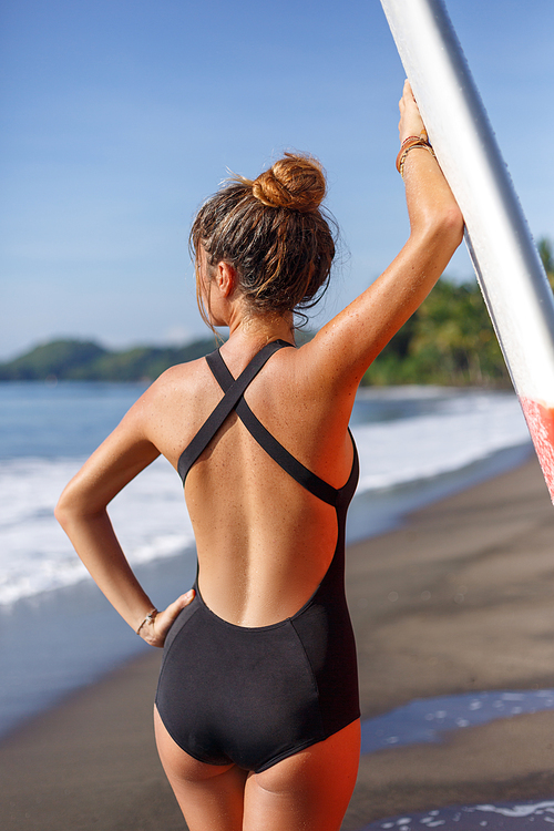 back view of young woman in swimsuit posing with surfboard near sea