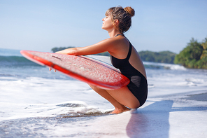 young woman in swimsuit sitting with surfboard near water