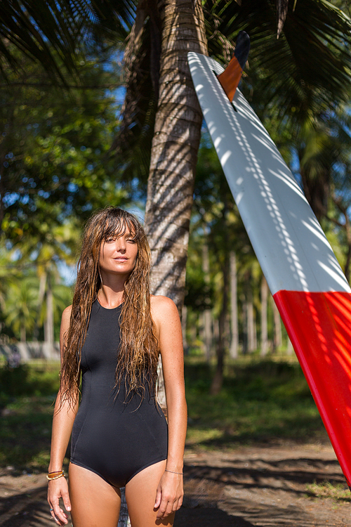 tanned girl with long hair standing with surfboard near palm tree