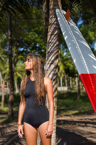 tanned girl in black swimsuit posing with surfboard near tree