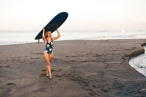 young sportswoman in swimming suit carrying blue surfing board on head on coastline